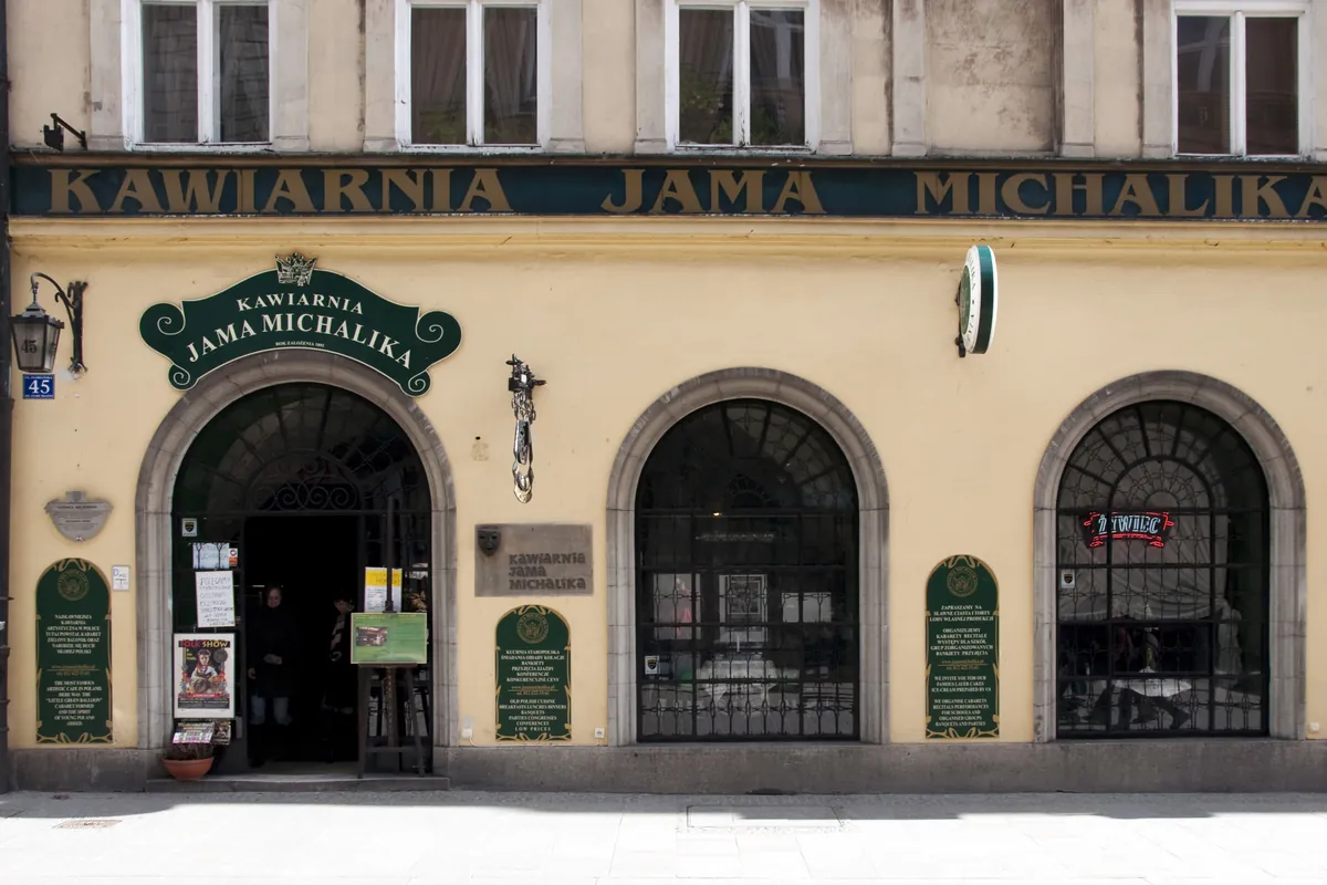 Żółta fasada Jamy Michalika przy ulicy Floriańskiej, z szyldem nad łukowym wejściem i dwoma łukowymi oknami z kratami.