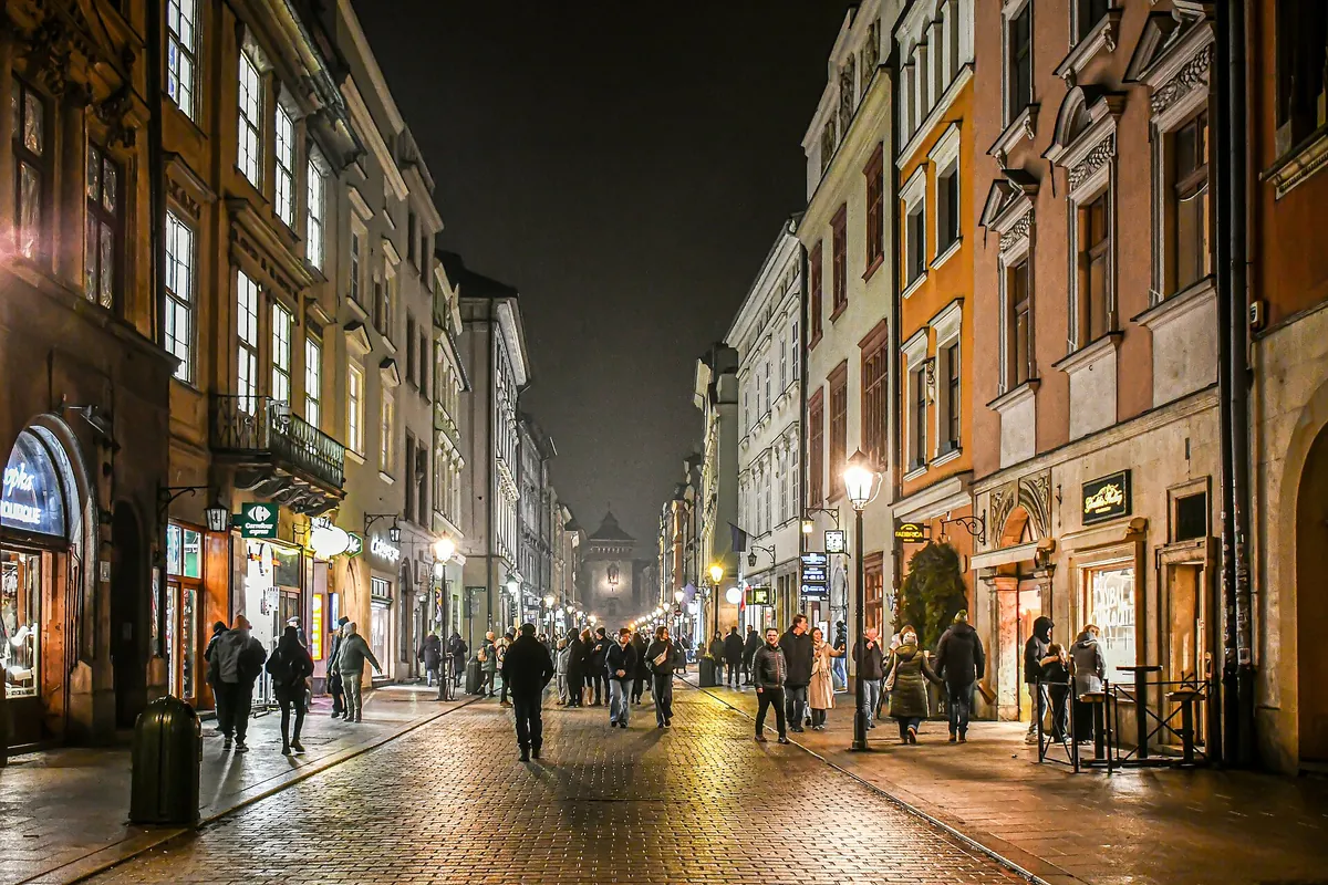 Ulica Floriańska nocą — brukowana ulica oświetlona latarniami i witrynami sklepów, z przechodniami i bramą miejską w oddali.
