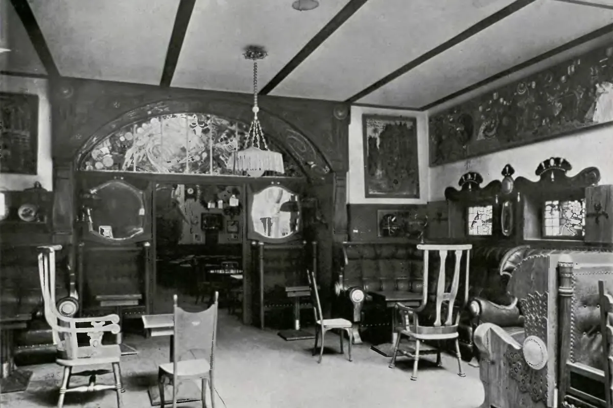 Černobílá fotografie interiéru kavárny z doby kolem roku 1911 s vitrážovým obloukem, kulatými zrcadly a světlými dřevěnými židlemi.