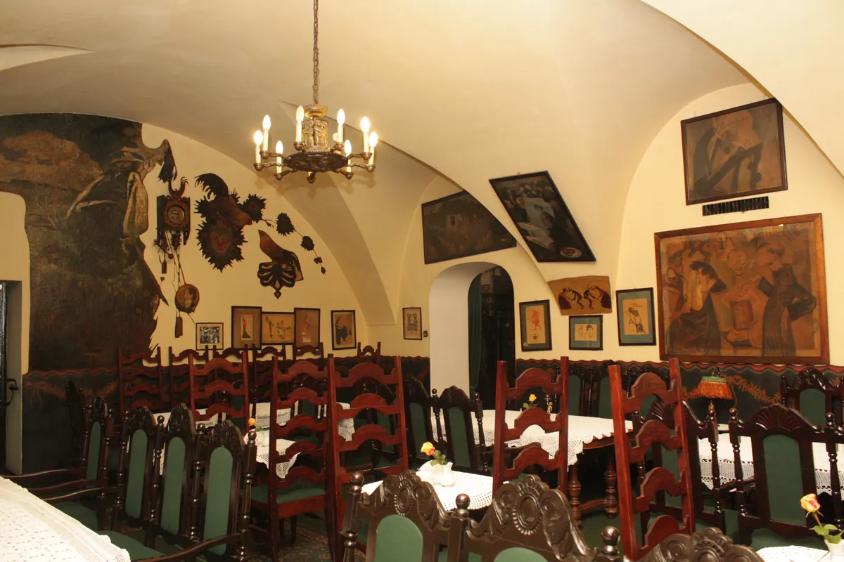 Sala ze sklepieniem, żyrandolem, oprawionymi karykaturami i obrazami na kremowych ścianach oraz ciemnymi drewnianymi krzesłami z zielonymi siedziskami.
